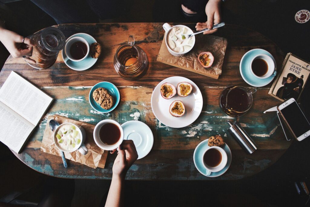 Top view of a table with coffee and pastries, with one hand reaching for a cup of coffee and another hand stirring a cereal bowl.
