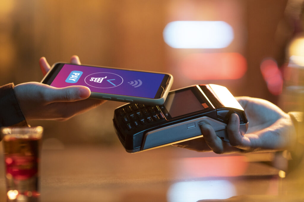 A customer making a contactless payment with a smartphone on a card reader machine while another hand holds the device.